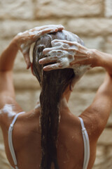 Woman washing hair with foamy shampoo, wet strands covered in bubbles, cleansing routine under warm...