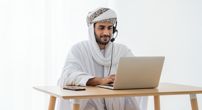 A man in traditional Middle Eastern attire uses a laptop and headset for a video call, working remotely from home.