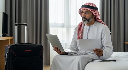 A businessman in traditional Middle Eastern attire sits on a hotel bed, working on his laptop and reviewing documents while his suitcase sits nearby.