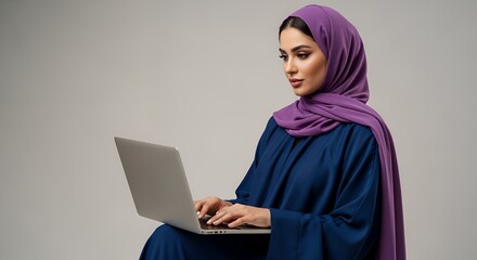 A young woman wearing a hijab sits and works on a laptop computer in a studio setting.