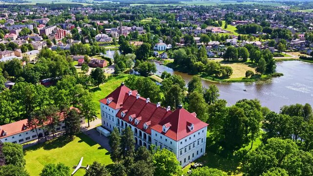 Aerial reveal shot of historic Biržai Castle with red roof, nestled in lush parkland beside a tranquil lake in Biržai, Lithuania.