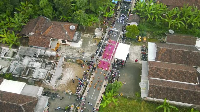 A festive street event with a stage, dancers, and a crowd in front of village houses and greenery.