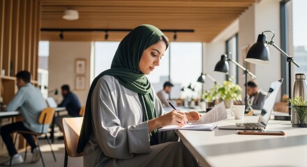 A Muslim woman in a hijab works diligently at her desk in a modern, sunlit co-working space, focused on her paperwork.