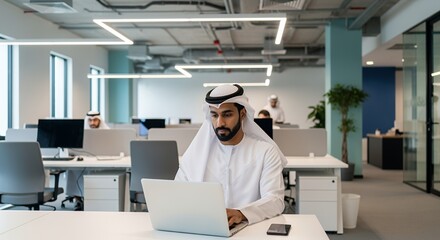 A young man in traditional Middle Eastern attire works diligently on his laptop in a modern, open-plan setting.