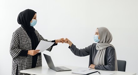 Two women in hijabs wearing face masks give each other a fist bump in a modern setting.