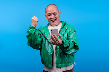 Senior Asian Man Cheering Excitedly While Looking At Smartphone Isolated On Blue Background