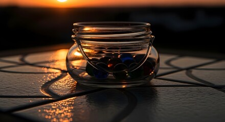 A glass jar filled with colorful marbles sits on a tiled surface during a vibrant sunset scene outside
