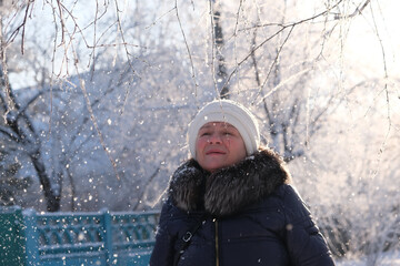 Woman in the blue padded jacket and white cap under snowfall at the street. Pensive attractive mature woman with a quiet smile. Sunny winter vacation outdoors. Deep breathing freshness
