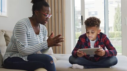A mother and son are sitting on the couch at home, the son is playing on a tablet, they are happy and enjoying their time together, they are a family and they love each other very much