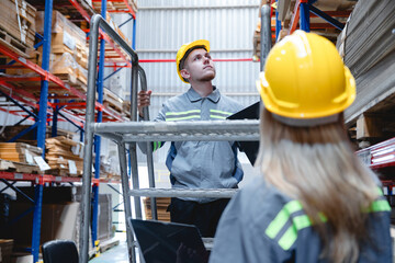 Warehouse workers in safety gear working as a team; male staff on ladder inspecting cardboard stock...