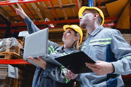 Two warehouse supervisors wearing safety uniforms using laptop and tablet to inspect cardboard stock on high shelves, showing smart logistics and teamwork.
