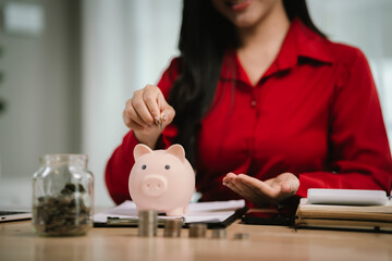 A smiling young woman sits at a desk indoors, holding a piggybank and coins, planning her finances...