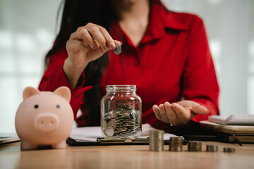 A smiling young woman sits at a desk indoors, holding a piggybank and coins, planning her finances with a calculator, symbolizing saving, security, and financial growth.