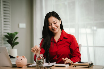 A smiling young woman sits at a desk indoors, holding a piggybank and coins, planning her finances...