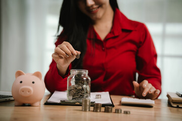 A smiling young woman sits at a desk indoors, holding a piggybank and coins, planning her finances...
