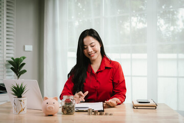 A smiling young woman sits at a desk indoors, holding a piggybank and coins, planning her finances...