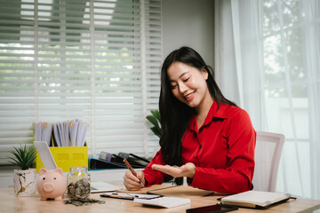 A smiling young woman sits at a desk indoors, holding a piggybank and coins, planning her finances...