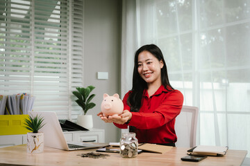 A smiling young woman sits at a desk indoors, holding a piggybank and coins, planning her finances...