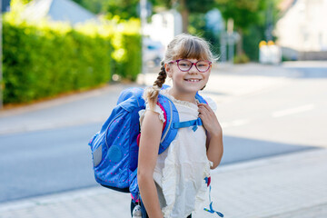 Cute little elementary school girl with eyeglasses walking to school in the morning with a backpack. Real life, real people, happy student on daily routine.