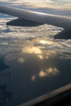 Fototapeta Golden sunset reflects over calm ocean waters and coastal land as seen from an airplane window, with the aircraft wing slicing across a sky filled with scattered clouds and warm hues.