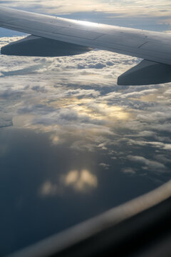 Fototapeta Golden sunset reflects over calm ocean waters and coastal land as seen from an airplane window, with the aircraft wing slicing across a sky filled with scattered clouds and warm hues.