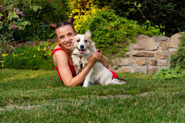 Woman sits on grass next to a happy white dog in a colorful garden surrounded by green plants and...