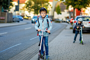 Two school kid boys in safety helmet riding with scooter in the city with backpack on sunny day. Happy children in colorful clothes biking on way to school.