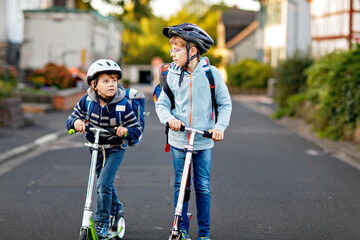 Two school kid boys in safety helmet riding with scooter in the city with backpack on sunny day. Happy children in colorful clothes biking on way to school.