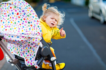Portrait of little sad toddler girl sitting in stroller and going for a walk. Crying baby child...