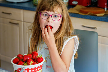 Little school girl with eyeglasses eating fresh sweet strawberries in home kitchen. Healthy food for children. Real life, real people, natural nutrition and fruit snack indoors.