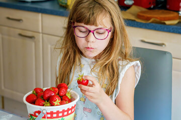 Little school girl with eyeglasses eating fresh sweet strawberries in home kitchen. Healthy food for children. Real life, real people, natural nutrition and fruit snack indoors.