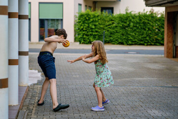 Little elementary girl and teenage boy, siblings, playing with ball in home garden on sunny summer day. Real life, real people, outdoor family fun, childhood bonding and joy