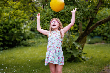 Little elementary school girl playing with a ball in home garden on a sunny summer day. Real life, real people, active outdoor childhood fun in domestic backyard.