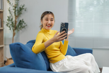 Obraz premium Happy young Asian woman making a video call with her smartphone while sitting on a comfortable blue sofa in her modern living room, enjoying a casual and connected lifestyle