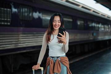 Smiling tourist is using mobile phone while standing with suitcase on platform at train station, checking online timetable, booking taxi, browsing internet or social media