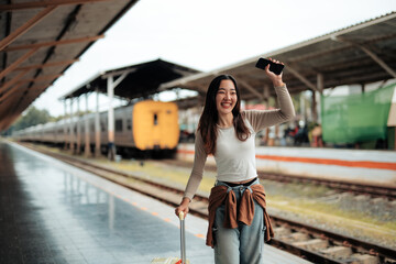 Smiling asian woman is waving her smartphone to call a taxi or greeting someone while pulling her trolley suitcase at train station after getting off the train