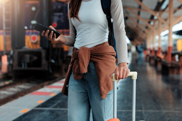 Young female tourist is standing on a train station platform, holding her trolley suitcase and...