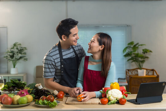Young happy asian couple wearing aprons is preparing healthy meal with various fresh colorful vegetables on kitchen counter at home, looking at each other, smiling and having fun - Powered by Adobe