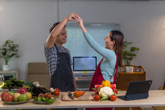 Happy Asian couple wearing aprons making high five gesture, celebrating successful meal preparation in modern kitchen with various fresh ingredients and tablet on the table