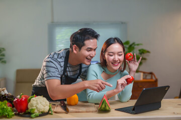 Young cheerful Asian couple wearing aprons having a video call on a tablet, showing and pointing at fresh tomatoes, with various fruits and vegetables lying on the kitchen table