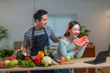 Young asian couple wearing aprons preparing healthy meal with lots of vegetables and having a video...