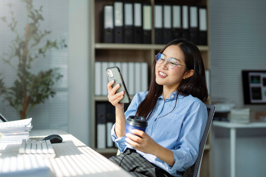 Smiling businesswoman wearing glasses, using a smartphone and holding takeaway coffee while browsing the internet during a break in a modern office - Powered by Adobe