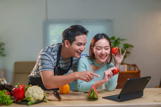Young cheerful Asian couple wearing aprons having a video call on a tablet, showing and pointing at fresh tomatoes, with various fruits and vegetables lying on the kitchen table