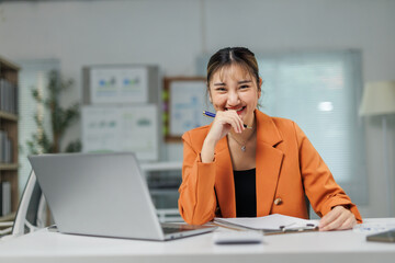 Happy young Asian businesswoman smiling while sitting at her desk, working on a laptop and...