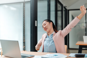 Young Asian businesswoman is stretching her arms while sitting at her desk in a modern office,...