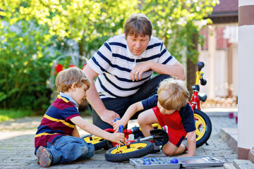 Two happy little kid boys and father repair chain on bikes and change wheel of balance bicycle. Family work together with working tools outdoors. Active leisure. Children, sons learning from dad