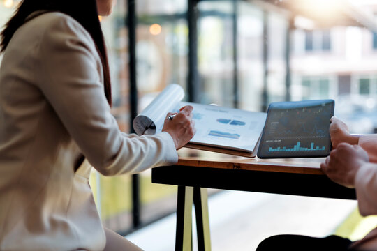 Two businesswomen discussing financial charts and graphs on a tablet and printed reports, collaborating on investment strategies in a modern office setting - Powered by Adobe