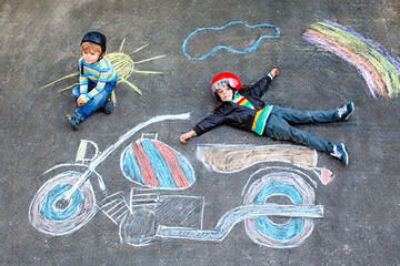 Two funny preschool boys wearing helmets drawing a motorcycle with colorful chalk outdoors. Real life, real people moment of creative leisure, friendship, and playful childhood fun