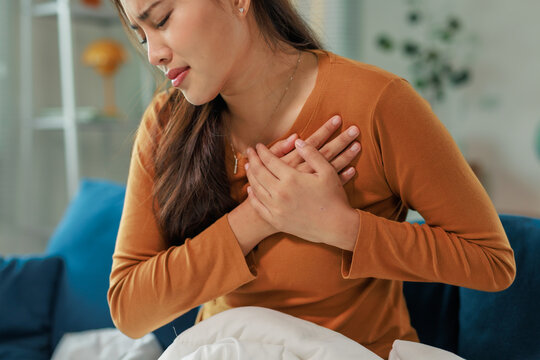 Young woman experiencing sudden chest pain, clutching her chest with both hands, her face expressing discomfort and worry, while sitting on bed in her home