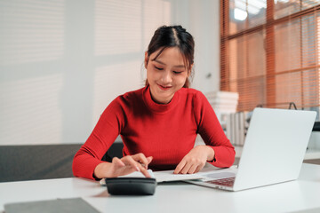 Young professional woman working at an office desk, using a calculator and laptop to analyze...